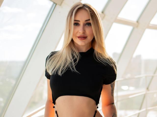 CandyGraces with blonde hair, blue eyes, and a cute smile, wearing a black crop top, showing her small tits and tattooed arm, standing in bright sunlight.