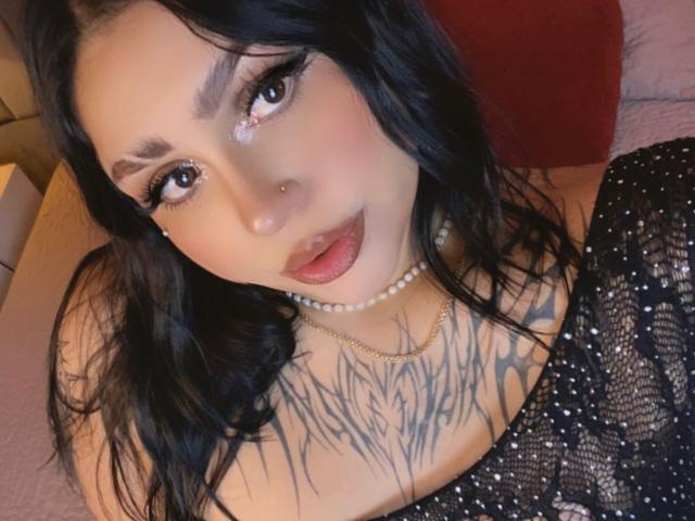 Emma Myles, avec ses cheveux foncés, son eye-liner et ses lèvres brillantes, portant un collier de perles et un haut noir scintillant, montrant un grand tatouage sur la poitrine et un piercing au nez, regardant l'appareil photo..