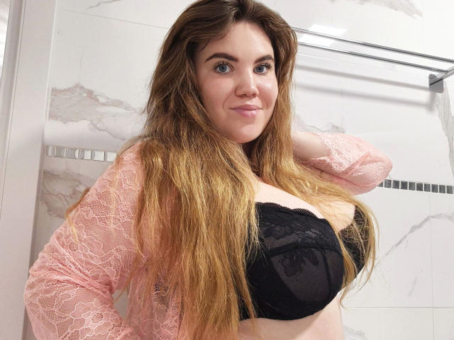 Mirabella4U-hot with long brown hair, wearing a black lace bra and pink lace top, smiling in a bathroom, showcasing her big tits.