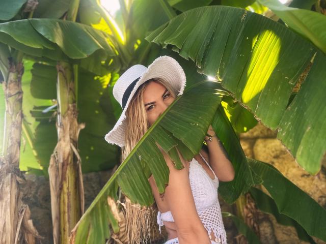 ZoeyMarie with blonde hair, wearing a white hat and mesh top, holding a large green banana leaf, her tits barely visible, surrounded by lush banana plants, sunlight filtering.