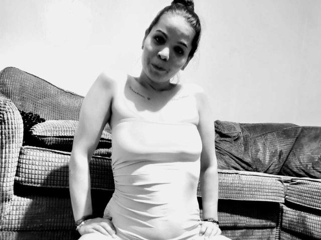 SweetLightWom ha dei piccoli seni, indossa una canottiera bianca aderente, è seduta su un divano a scacchi con la testa leggermente inclinata e un piccolo sorriso, in una foto in bianco e nero..