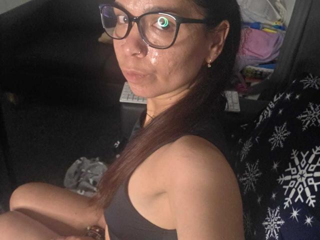 SweetLightWom with shiny face, green-eyed glasses, black tank top, and brown hair, sitting on a black and white snowflake-patterned chair, looking at the camera with a pouty expression.