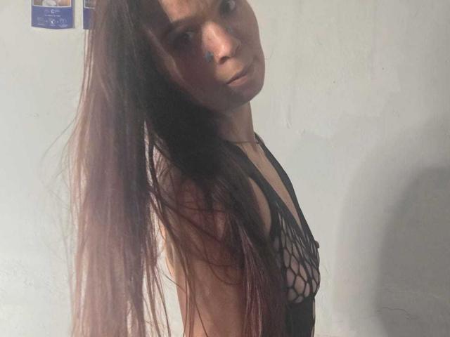 SweetLightWom with long brown hair, wearing a black netted top that reveals her small tits and a blue face paint mark, looking slightly to the side.