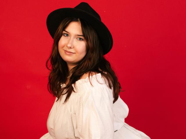 TrishaKingsley with wavy brown hair, wearing a black hat and off-shoulder white dress, has a cute smile, and is posing against a bright red background.