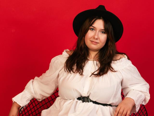 TrishaKingsley with brown hair, wearing a black hat and white dress with a black belt, sitting on a red chair, smiling at the camera. Boobs, cute.