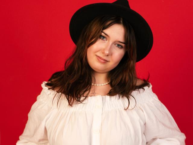TrishaKingsley has blue eyes, wavy brown hair, a black hat, a white off-shoulder shirt, and a pearl necklace, with a slight smile against a bright red background.