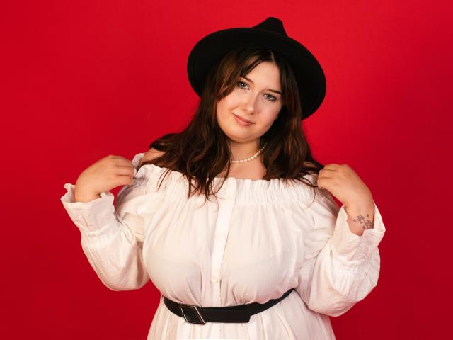 TrishaKingsley with big boobs, wearing a white dress, black belt, and black hat, holding her dress up, smiling, red background, brown wavy hair, pearl necklace, tattoo on wrist.