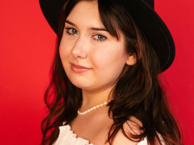 TrishaKingsley with wavy brown hair, wearing a black hat and white dress, pearl necklace, and subtle smile, against a red background, showing cute green eyes.