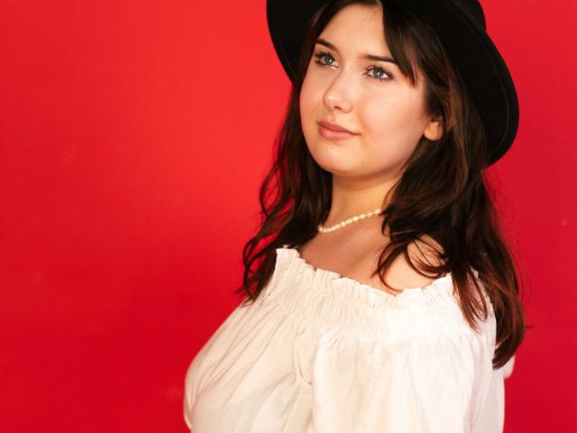 TrishaKingsley with wavy brown hair, wearing a black hat, white off-shoulder top, and pearl necklace, looking at the camera with a subtle smile, against a bright red background.