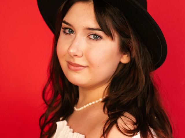 TrishaKingsley with wavy brown hair, wearing a black hat, white blouse, and pearl necklace, has green eyes and a subtle smile, against a bright red background.