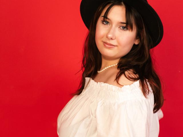 TrishaKingsley with brown hair, wearing a black hat, white off-shoulder top, and pearl necklace, has a cute smile and looks at the camera. Red background.