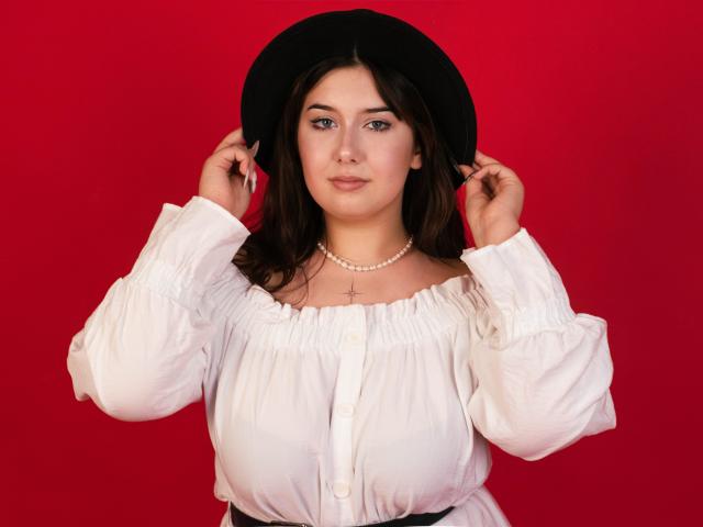 TrishaKingsley with medium tits, wearing a white blouse, black hat, pearl necklace, and holding her hat, against a red background. She has brown hair and a cross necklace.