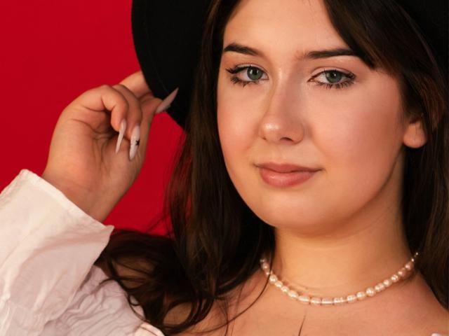 TrishaKingsley with a black hat, white shirt, pearl necklace, and long brown hair, holding her hat against a red background, showing her tits, cute face, and green eyes.