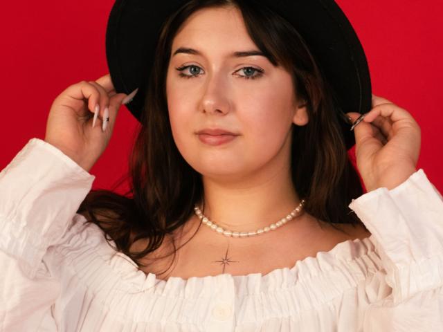 TrishaKingsley with dark hair, wearing a black hat, white ruffled shirt, pearl necklace, and a cross necklace, holding the hat with both hands. Red background.