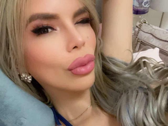 HotCumAdriana69x with pouty pink lips, winking, and blonde hair, wearing blue, with one arm raised, showing off a cute face and sexy earrings.