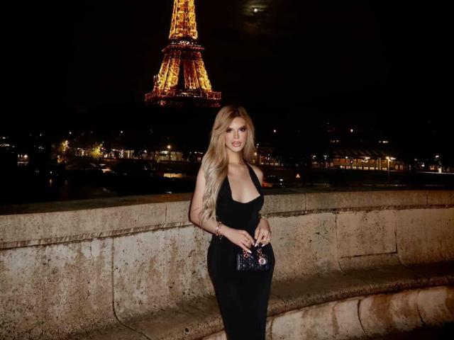 HotCumAdriana69x with long blonde hair, black dress, and cleavage stands by a stone wall, holding a small purse, with the Eiffel Tower brightly lit in the night.