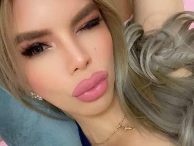 HotCumAdriana69x has pouty pink lips, long blonde hair with gray highlights, and thick, sexy eyebrows. She's winking with heavy eyelashes and a slight smirk.
