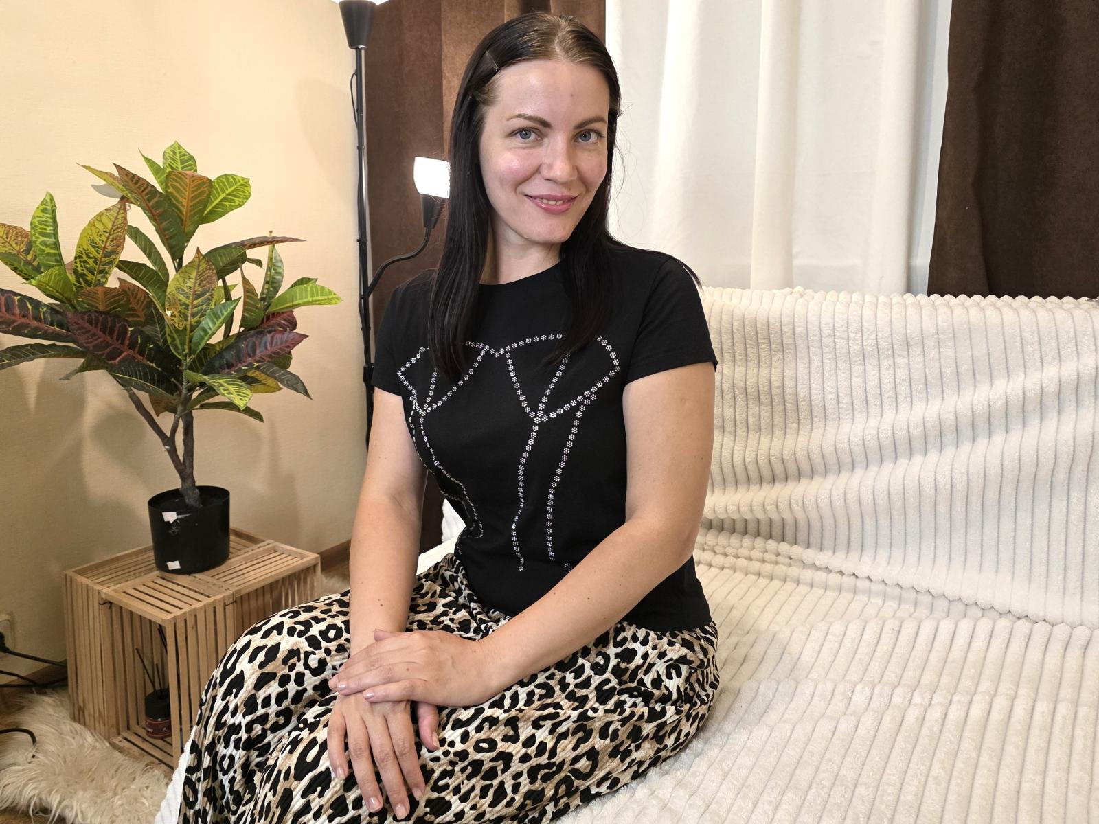 SaraLoritte with black top, leopard print pants, and white textured blanket, sitting on couch, smiling, hands on knees, potted plant to left, standing lamp behind.