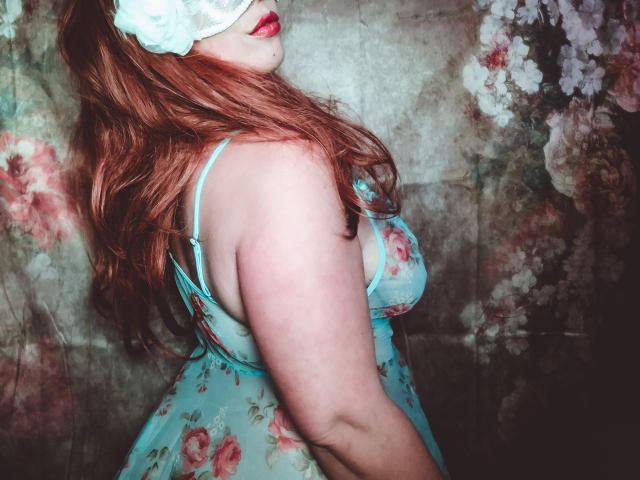 JuliaFrancaise, con i capelli rossi, indossa un abito intimo floreale blu, una benda per gli occhi bianca e rossetto rosso, mostrando la schiena e i glutei. Il suo seno è parzialmente visibile..