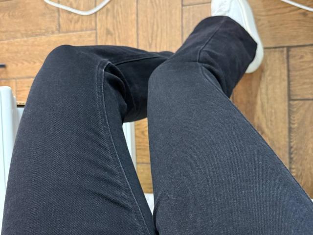Le jean noir, légèrement usé, et les baskets blanches d'Amabel, les jambes croisées, révèlent la texture du denim et le motif du plancher en bois. Vue de dessous, avec un focus sur les jambes..