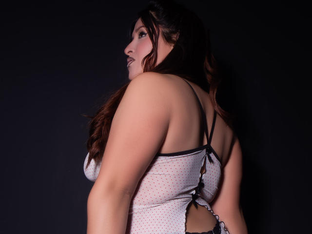 TaylorBecker with long brown hair, facing side, wearing a white polka dot lingerie top, black straps, cut-out back, showing her bare ass, against a dark background.