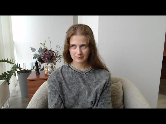 LariStella with long brown hair, blue eyes, and gray sweater, sitting in a beige chair, looking at camera, small potted plant and vase in background.