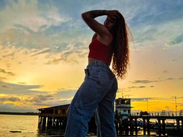 Valery Hilton mit langem, gewelltem Haar, die ein rotes Crop-Top und blaue Jeans mit hoher Taille trägt, steht auf einem Steg, mit dem Rücken zur Kamera, die Hände im Haar, Sonnenuntergang..