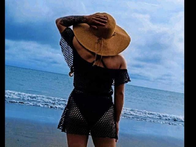 ValeryHilton stands on the beach, wearing a black mesh dress, off-shoulder top, and a wide-brimmed straw hat, covering her tits with her hand. Ocean and clouds behind.