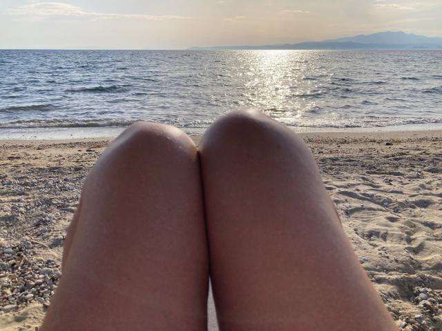 DeesseBleue's knees are in the foreground, with her bare thighs visible. She is sitting on a sandy beach, facing a shimmering ocean under a partly cloudy sky.