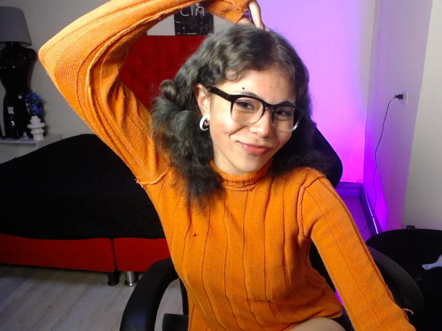 BeautyInsomnia with curly black hair, wearing an orange rib-knit sweater, black glasses, and white ear gauges, raising her arm, smiling, showing a cute face and small nose.