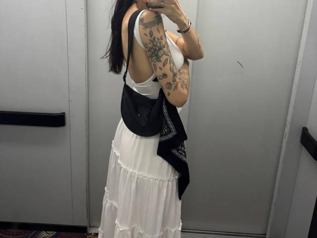 Tsukerberg con tatuajes en el brazo, vestida con un vestido blanco con volantes, una camiseta negra sin mangas, un bolso negro, sosteniendo un teléfono en un espejo, de pie en un ascensor..