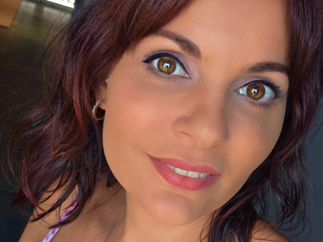 Close-up photo of JuneLaFrenchie, a brunette woman with brown eyes, wearing pink lipstick and a purple top, looking directly at the camera with a slight smile. Focused on her face, highlighting her expressive eyes and smooth skin.