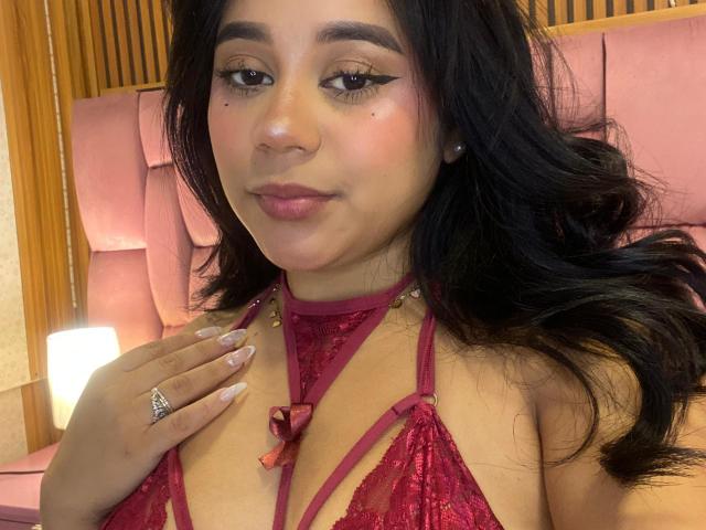 AuroraDiMarco with long black hair, wearing red lace lingerie, showing her small tits, a diamond ring on her finger, and pink nail polish, smiling softly.