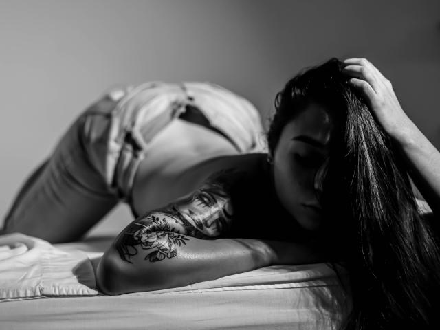 Black and white photo of AlahiaRouse on a bed, butt up, wearing unbuttoned jeans, arms tattooed, long dark hair, hand in hair, looking down, butthole visible, ass raised.
