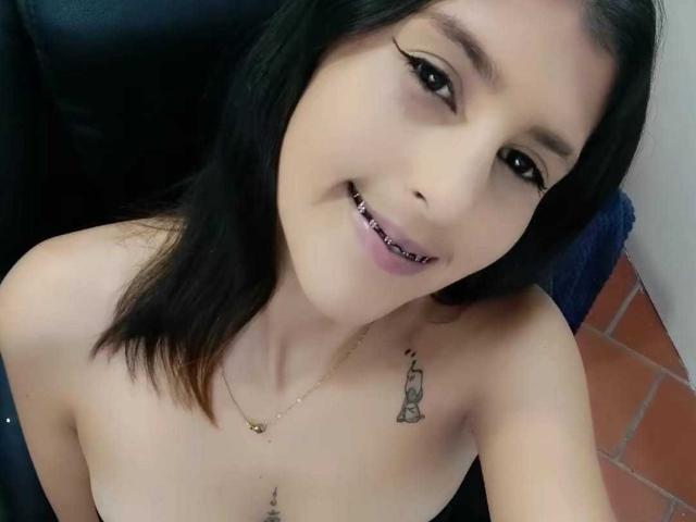 AlanaSexx with black hair, smiling, showing pierced teeth, gold necklace, small tattoo on right shoulder, and topless, with visible small tits and a nipple piercing.