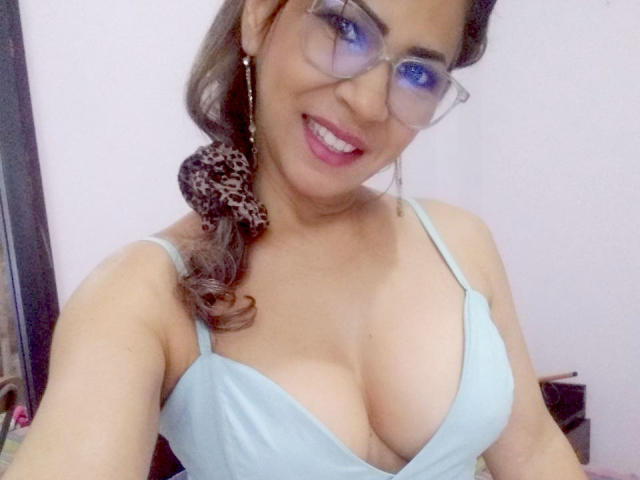 BrigiteWalsh with glasses, smiling, wearing a light blue tank top that shows her big, shiny tits. Brown hair in a ponytail, pink lipstick, and a leopard print scrunchie.