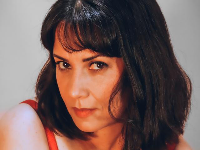 NectarAgave ha capelli scuri e ondulati e un'espressione seria. Indossa una maglietta rossa e i suoi occhi marroni guardano leggermente di lato. Il suo seno è parzialmente visibile..