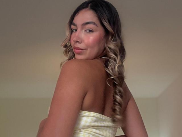 MaraCortez with wavy blonde and brown hair, looking over her shoulder, wearing a strapless, shiny yellow top, showing her back and slightly turning to show her tits.