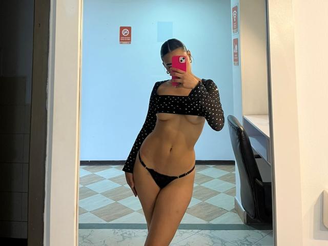 SienaReign with a slim, toned body, small bare tits, wearing a black polka-dot crop top and black panties, taking a mirror selfie in a room.