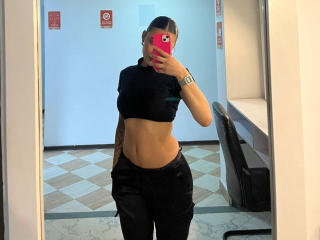 SienaReign with a slim, toned midriff, black crop top, black pants, and a tattoo on her right arm, taking a mirror selfie, holding a pink phone.
