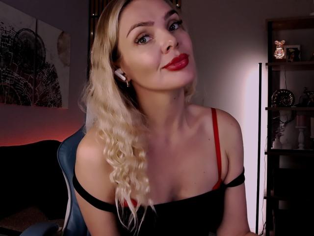 MoniDream with blonde curly hair, red lipstick, black off-shoulder top, red bra strap, white earbuds, smiling, light skin, blue eyes, sitting in a dimly lit room.