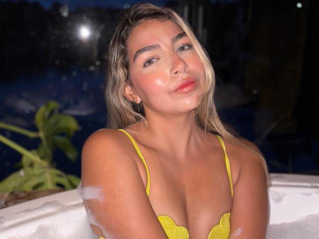 MaraCortez with wet blonde hair, yellow lace bra, and soap bubbles on her chest, pouty lips, and blue eyes, in a dark, nighttime setting, looking at the camera.