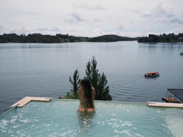 MaraCortez, back to camera, in a blue-tinted hot tub, water up to her ass, long brown hair, lake in the background, small boat on the right, overcast sky.