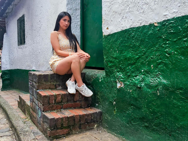 JainaRogers, con cabello negro largo, vestida con un vestido amarillo brillante y zapatillas blancas, sentada en escalones de ladrillo, con una pared verde y blanca detrás de ella, piernas cruzadas, manos sobre las rodillas..
