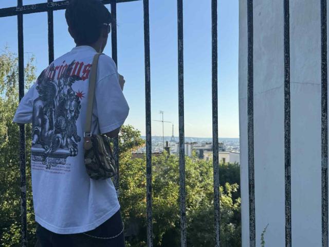 WillStarr with dark hair, wearing a white shirt with a graphic, stands by a black metal fence, looking outside at a clear blue sky and distant buildings.