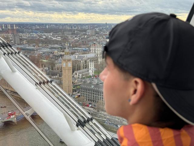 WillStarr's face with a black cap, orange and red striped shirt, and blue ear piercing, looking at London's skyline with the Tower Bridge and Thames River in the background.