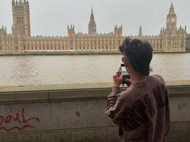 WillStarr with short brown hair, wearing a brown sweatshirt, stands by a river, holding a smartphone, looking at the camera, in front of the ornate, historic, London building.