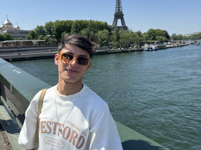 WillStarr with short brown hair, orange sunglasses, white Estford shirt, smiling, standing by a river, Eiffel Tower in background, clear blue sky, green trees, sunny day, casual vibe.