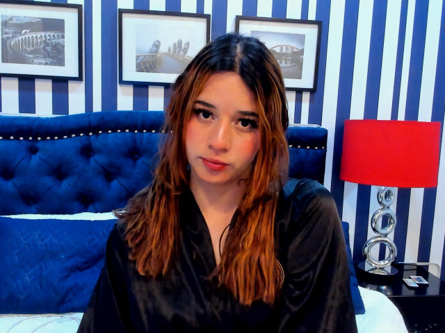 DaniVelasquez with wavy brown hair, wearing a black satin robe, sitting on a blue tufted bed, red lamp, and striped wall, looking at the camera with a subtle smile.