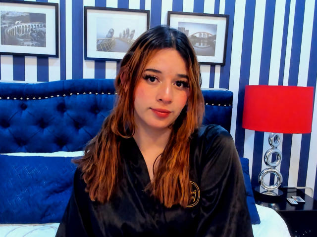 DaniVelasquez, com cabelos castanhos ondulados, vestindo um roupão de cetim preto, está sentada em uma cama azul com detalhes em botão, com uma luminária vermelha e um fundo de parede listrado. Ela tem uma expressão adorável, ligeiramente satisfeita..